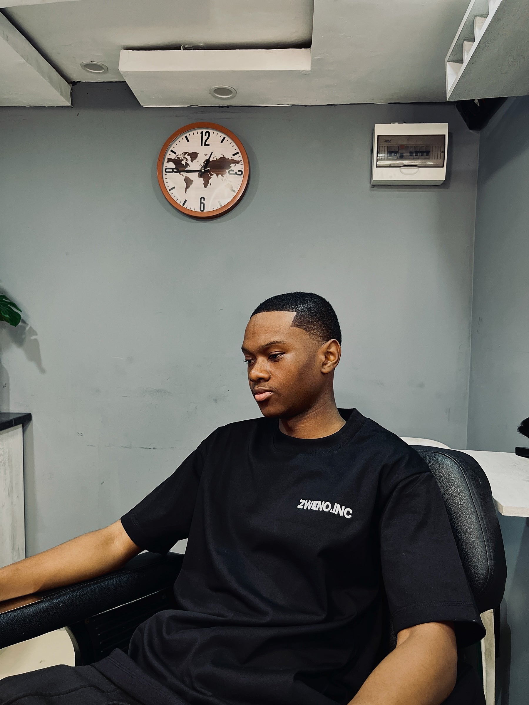 A man wearing a black ZWENO.INC cotton t-shirt with a minimalist white logo on the chest in a modern indoor setting.